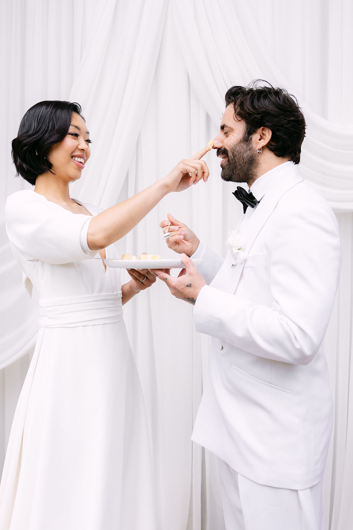 Bride touching cake to the groom's nose, both smiling, plates in hand