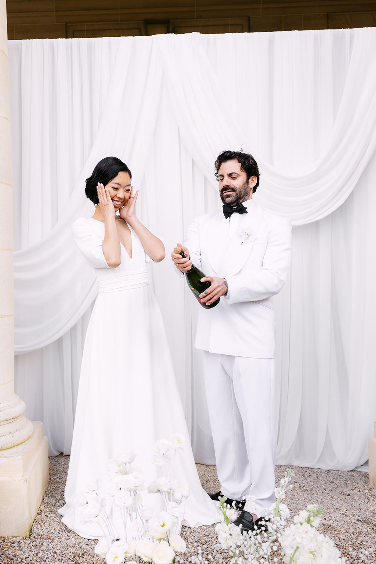 The bride covers her face laughing while the groom, in a white suit, opens a bottle of champagne before white drapes.