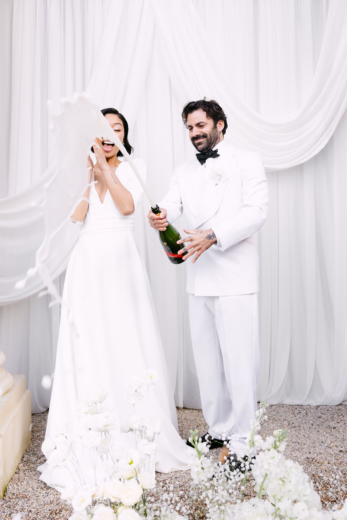 The groom holds a champagne bottle and laughs while the bride covers her face, both dressed in white before draped fabric.