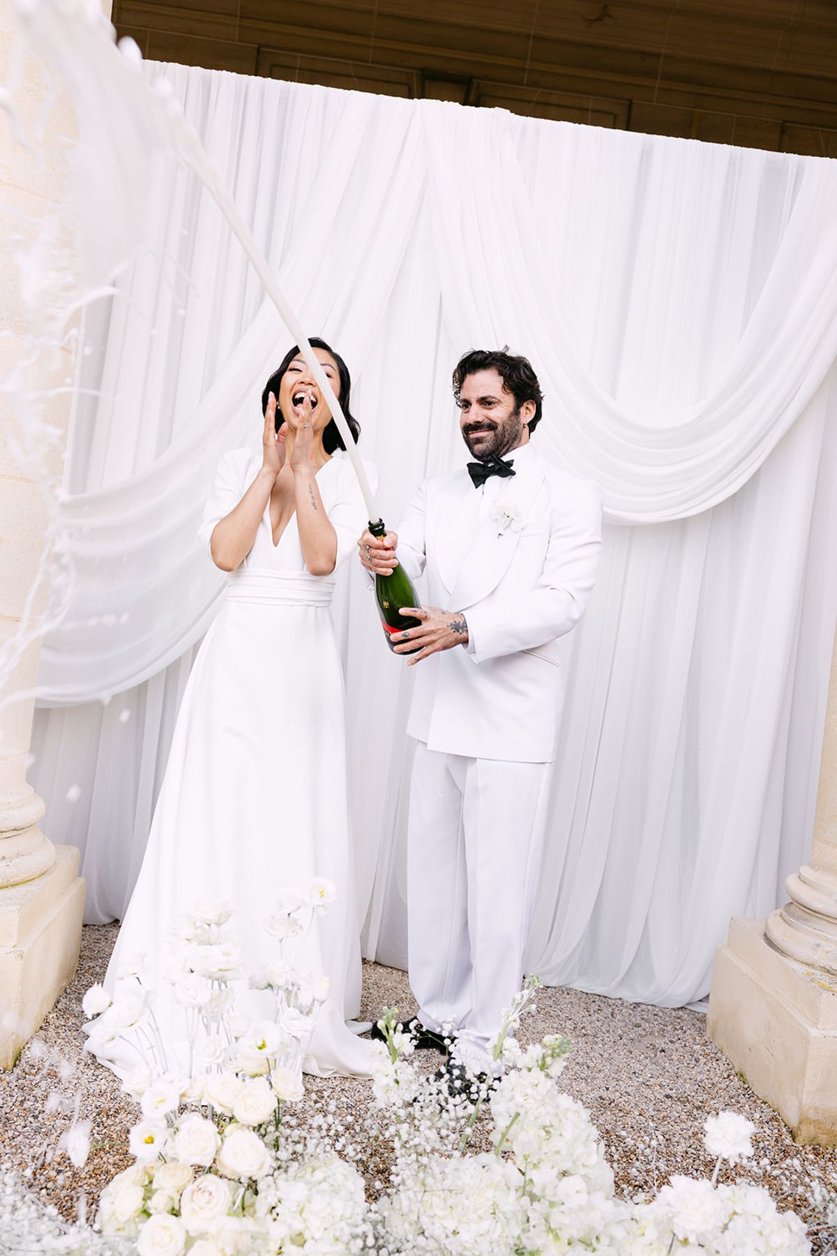 The couple in white stand before white drapes as the groom holds a champagne bottle and the bride raises her hands as it sprays.