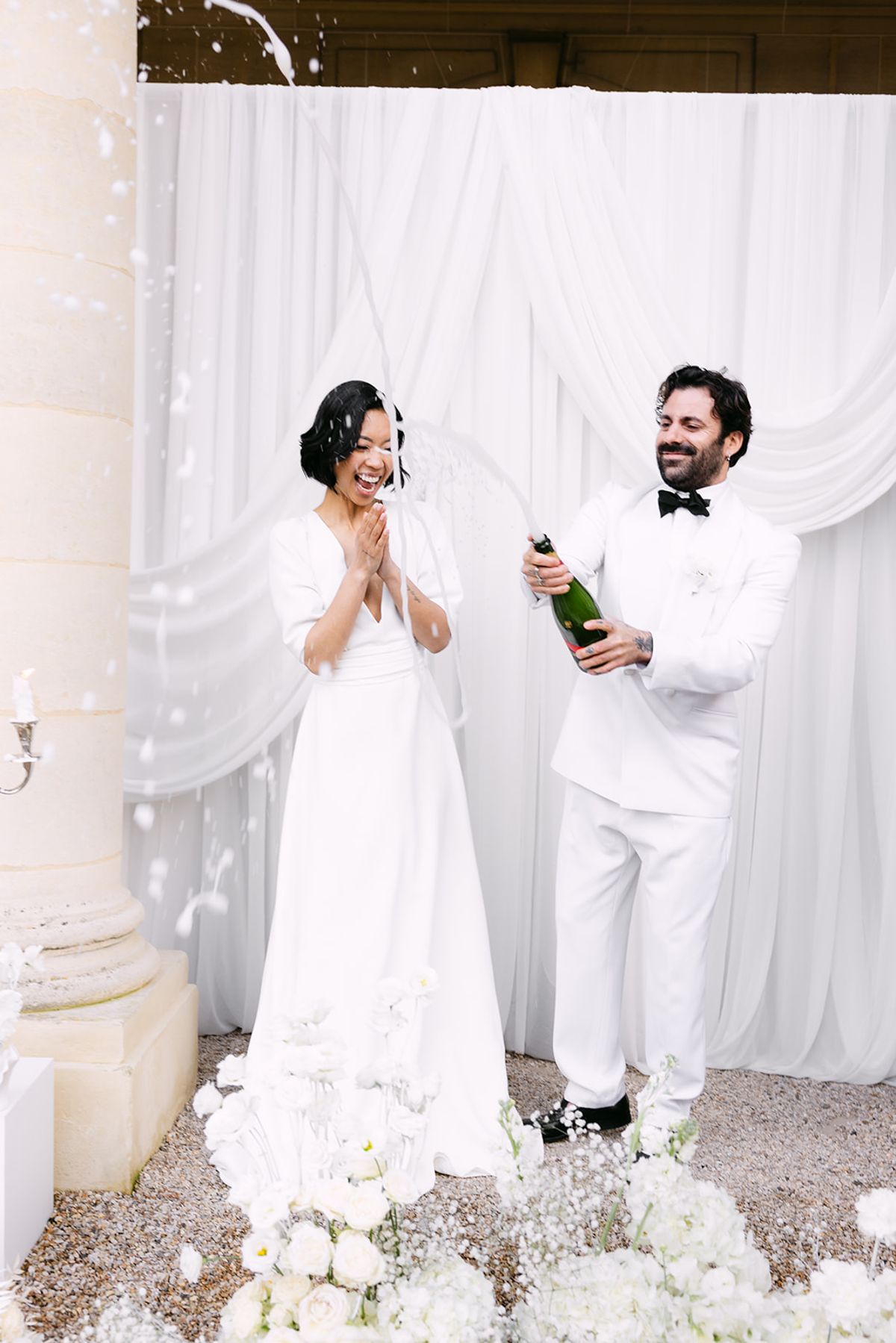 The bride claps as champagne sprays while the groom holds the bottle, both in white before draped fabric and a column.