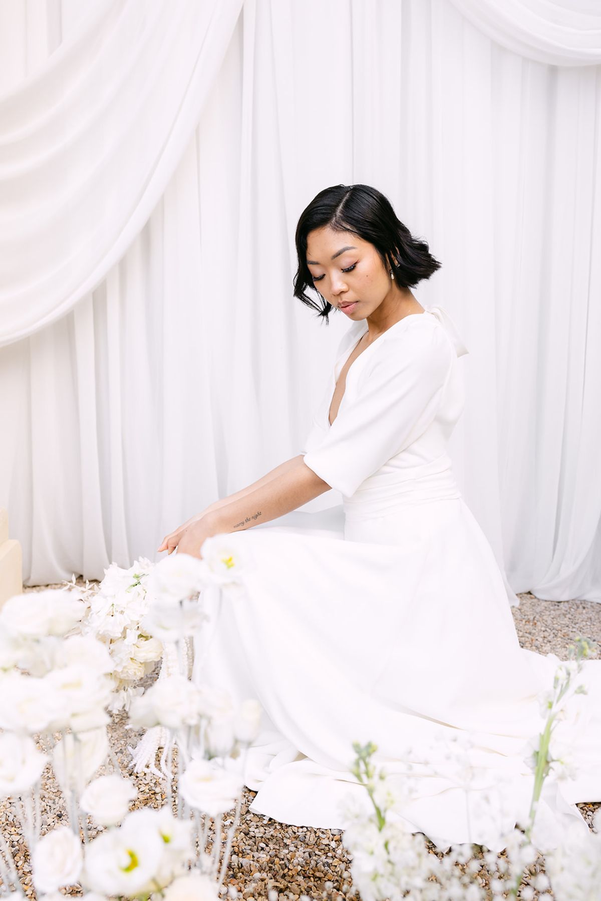 The bride in a white v-neck dress sits among white flowers before white drapes, looking down with her dark hair in a bob.