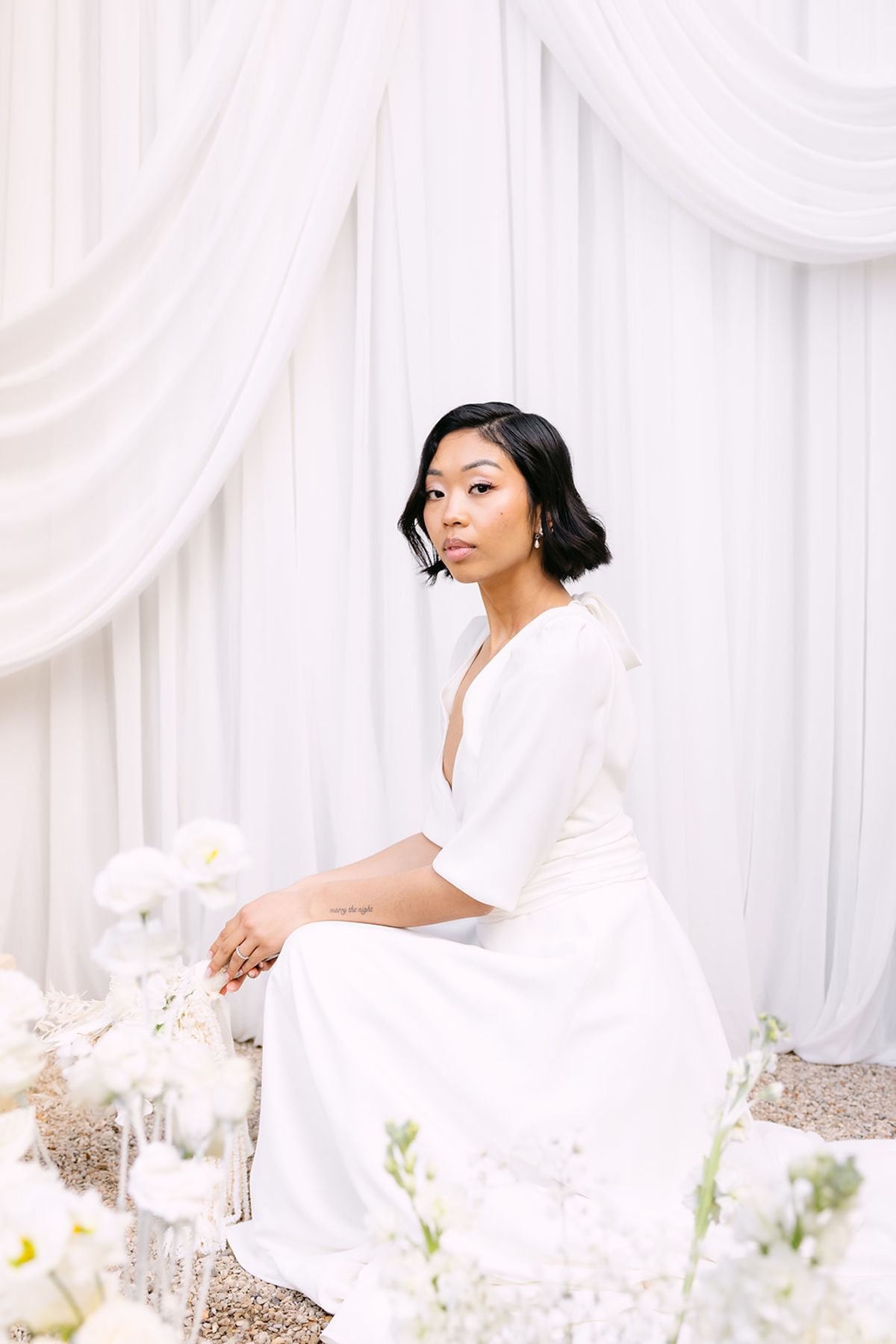 The bride in a white dress sits among white flowers before white drapes, looking towards the camera with a calm expression.