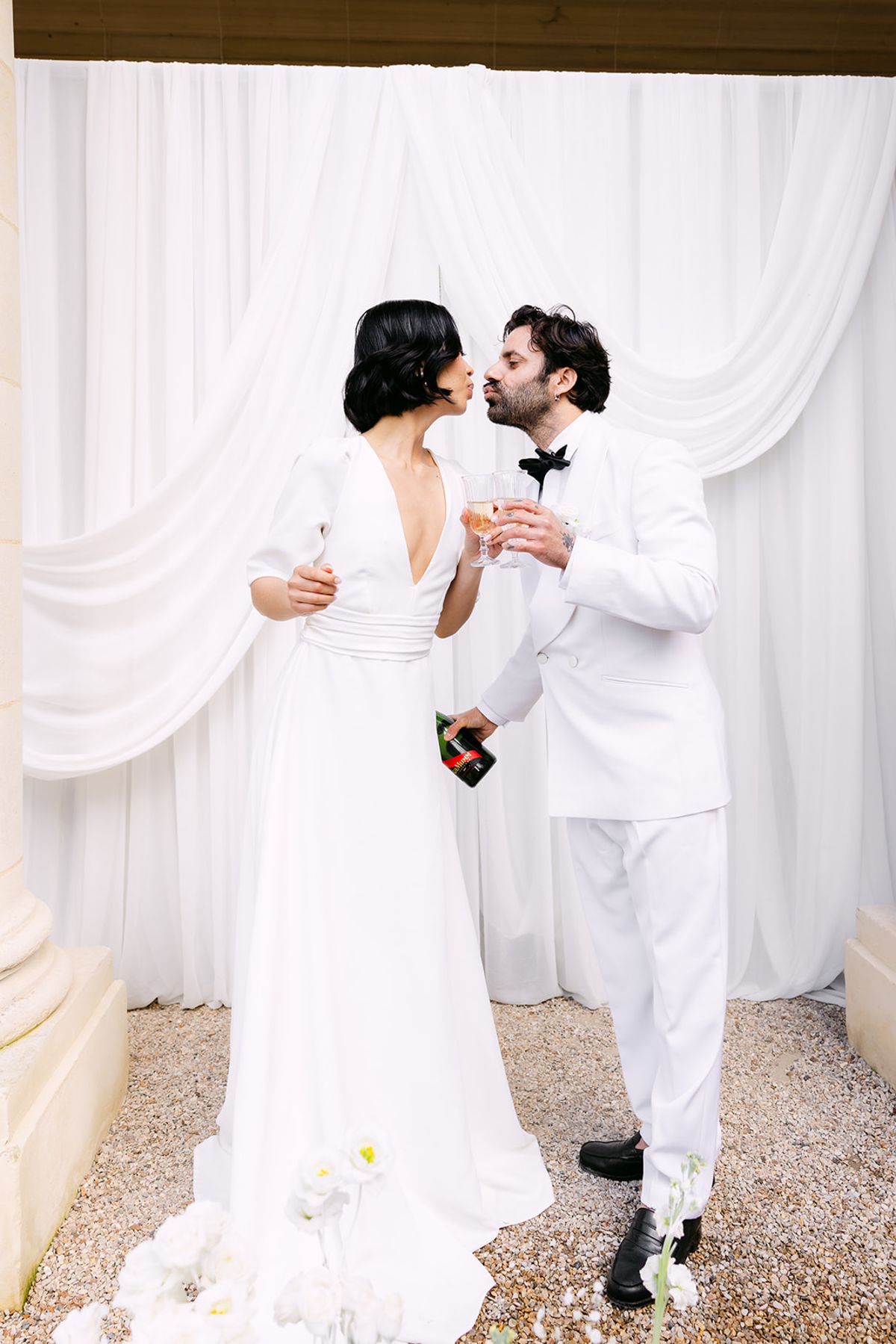The groom kisses the bride's cheek as they hold champagne glasses before white drapes, a bottle in his hand.