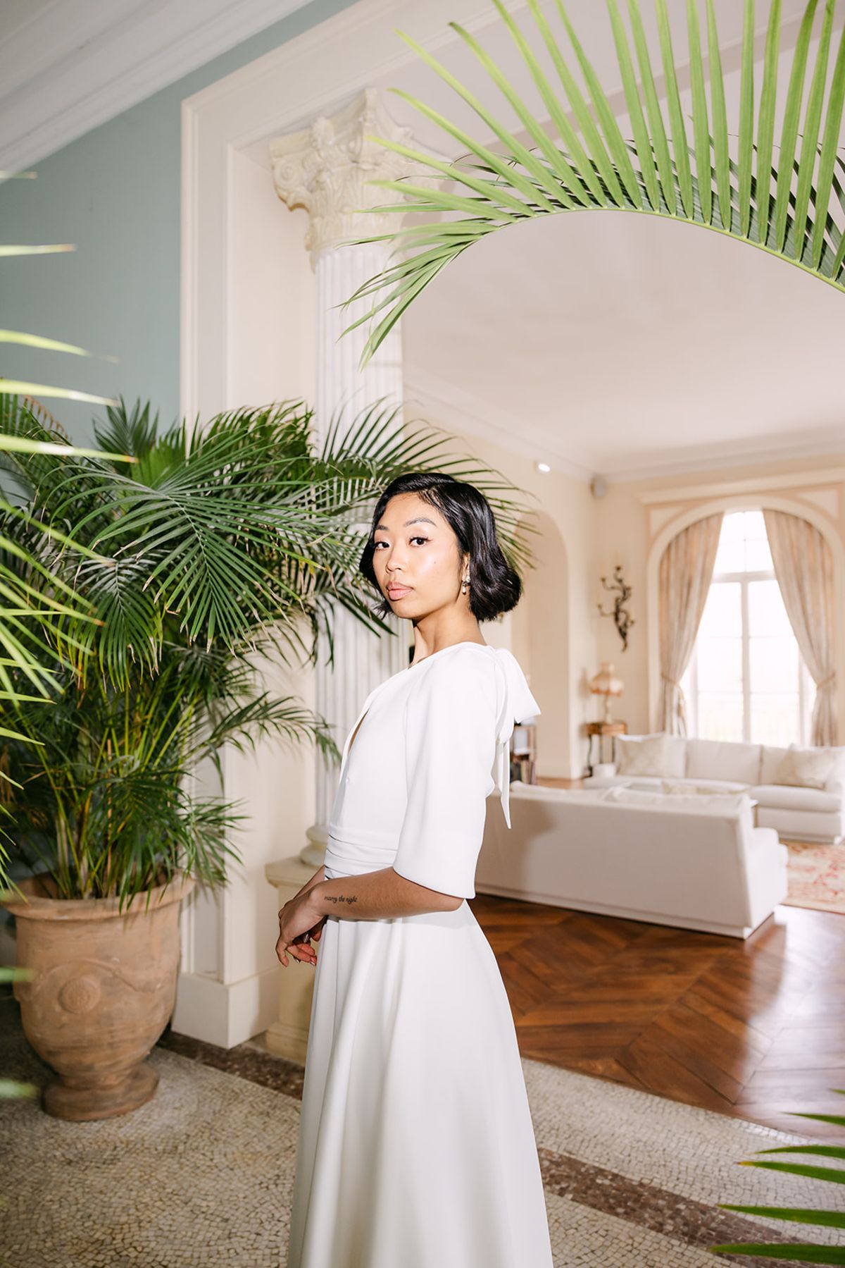 The bride in a white top and long skirt stands among palm plants in a bright room, looking towards the camera.