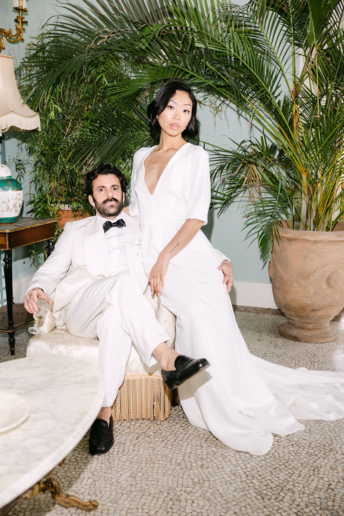 The groom sits in a white suit on a chair while the bride perches on its arm beside a large potted palm.