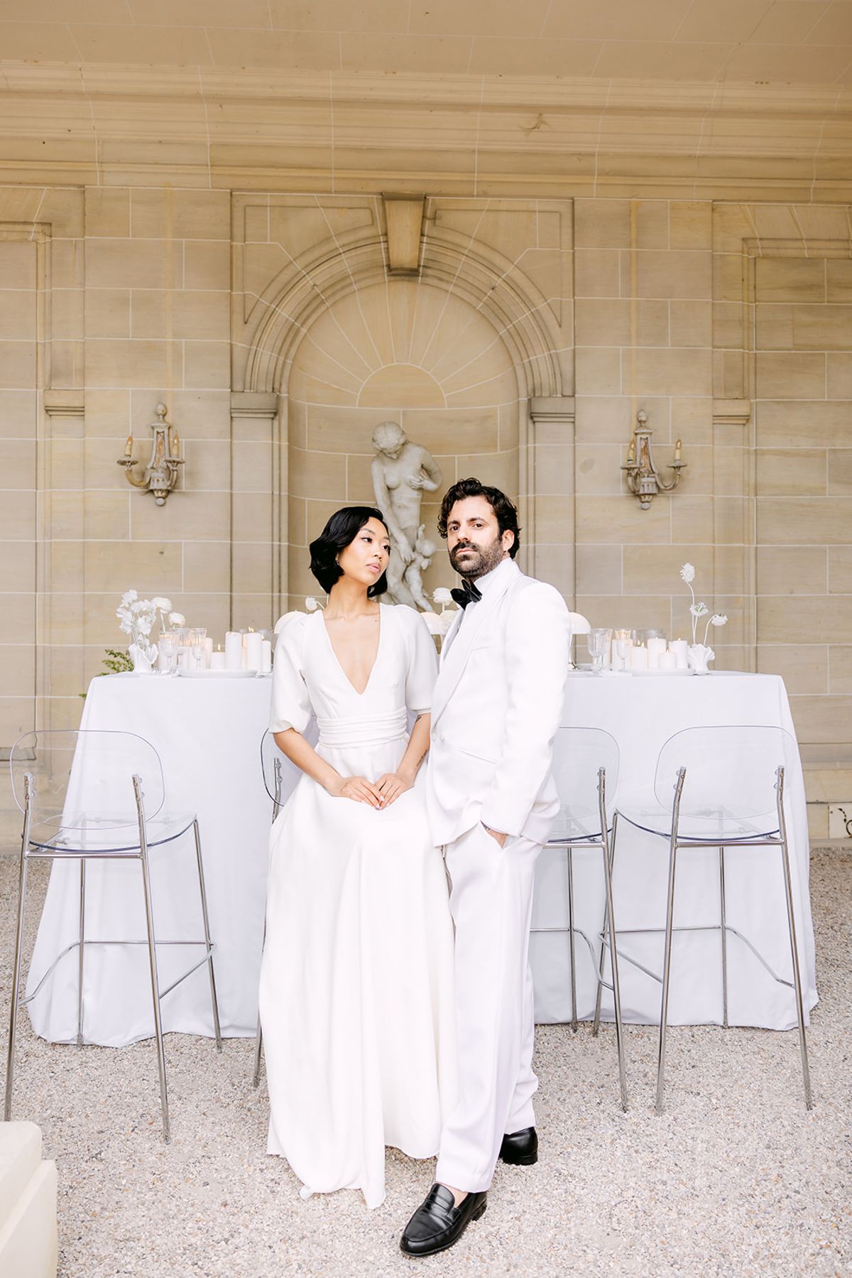 The bride and groom, both dressed in white, stand side by side before a white-clothed table and stools beneath a stone alcove with a statue.