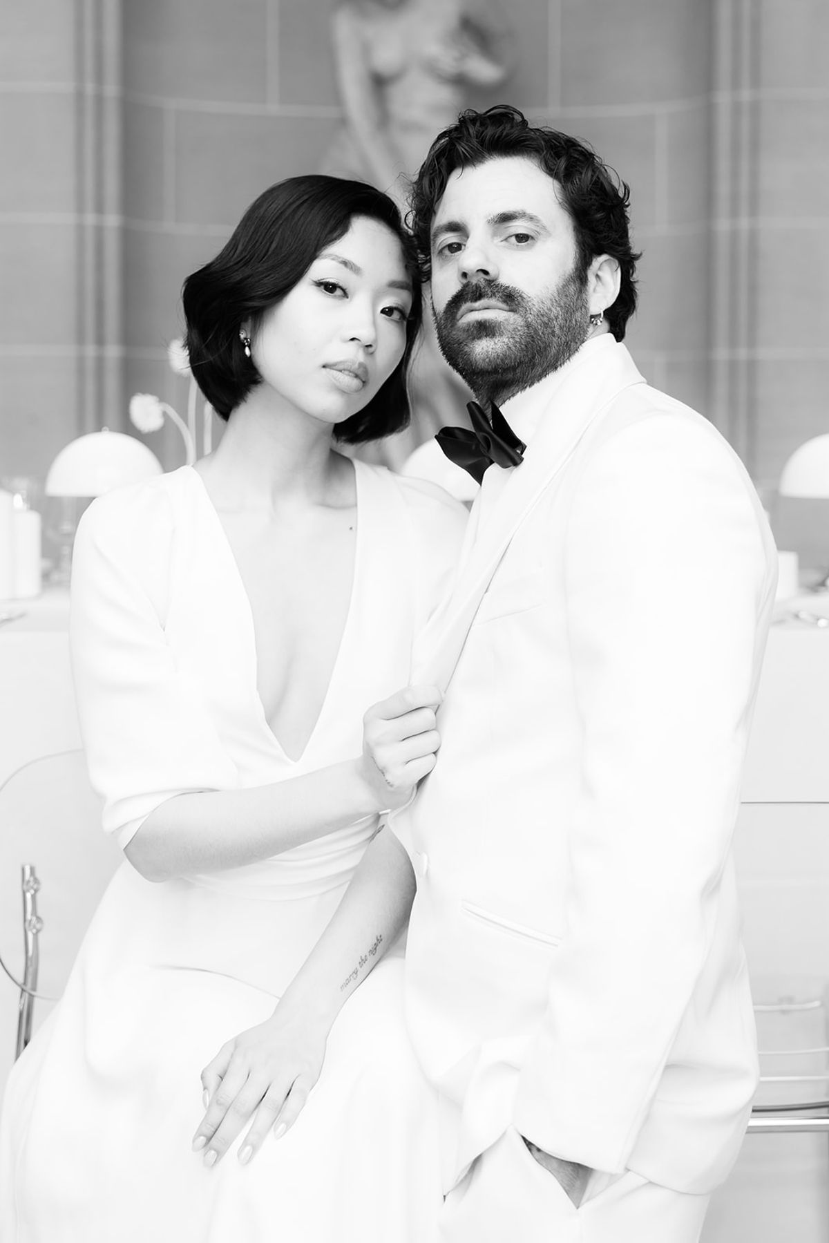 In a black and white portrait, the bride and groom sit close together in white outfits, looking towards the camera.