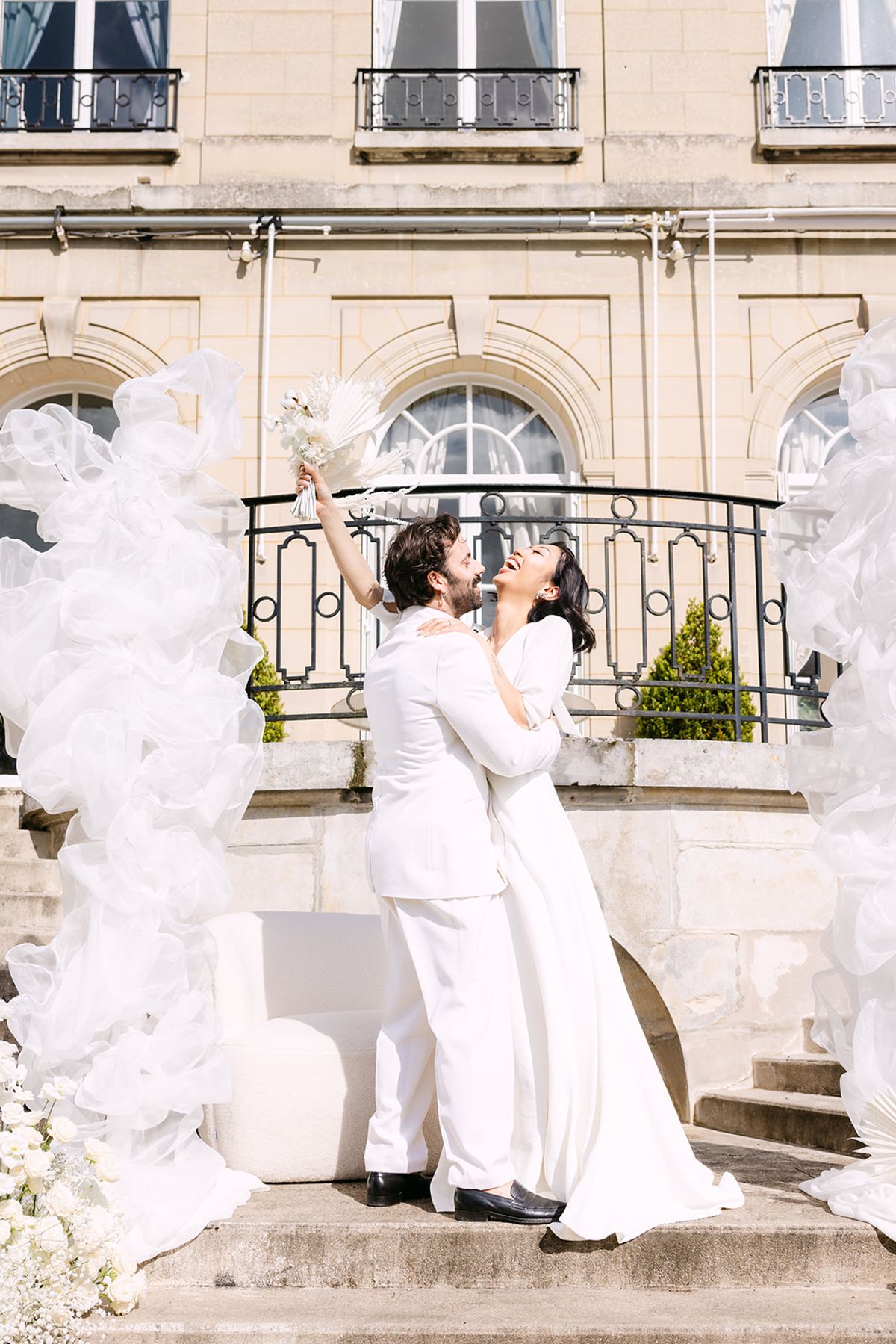 The couple in white embrace on stone steps before a chateau balcony as the groom lifts a small bouquet high.