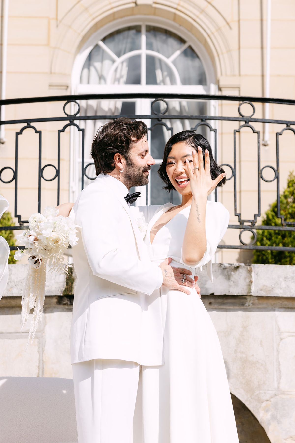The groom in a white jacket and bow tie holds the bride close as she laughs, hand to her face, before a balcony.
