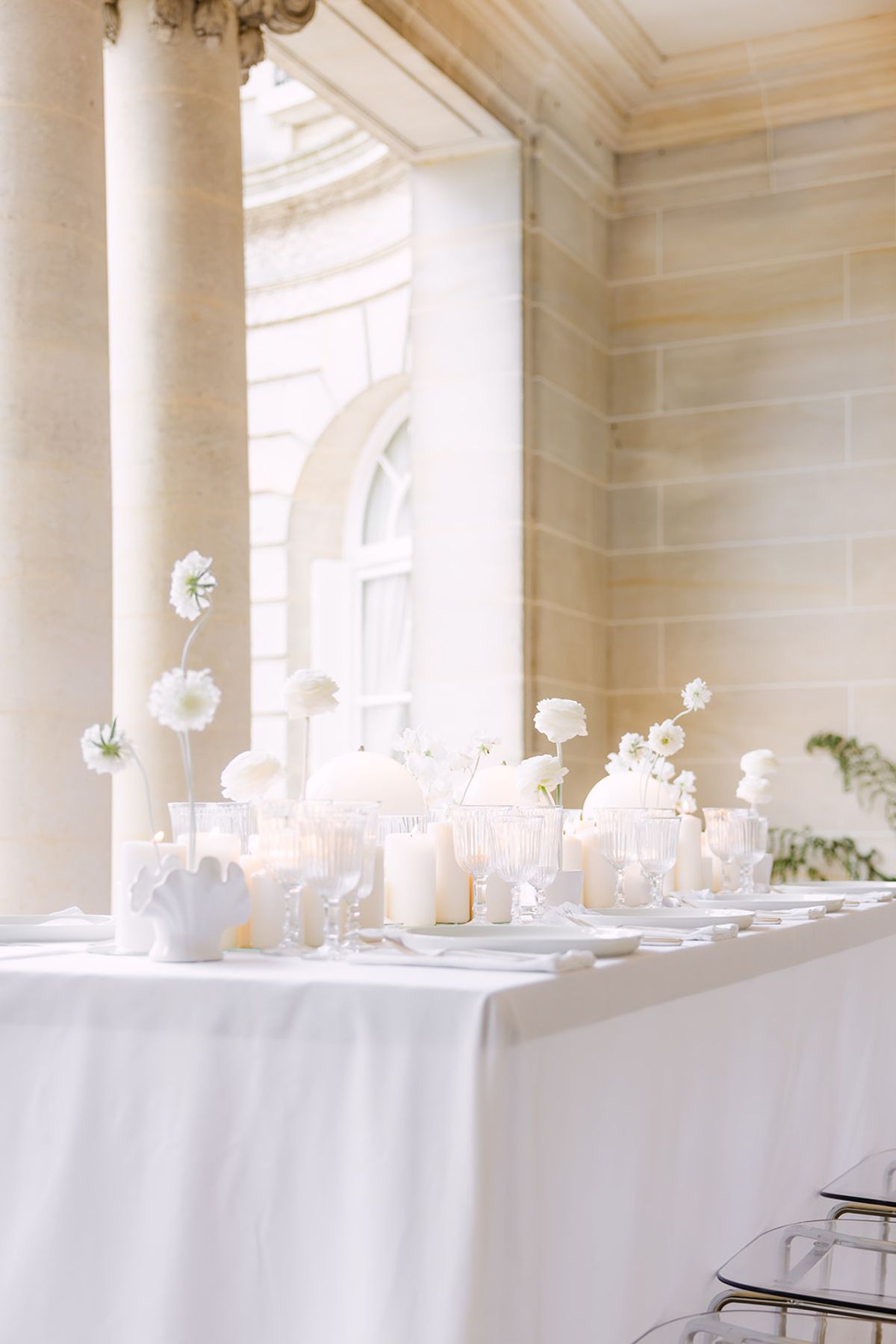 A long table with a white cloth is set with candles, small vases and white flowers in a stone columned gallery.