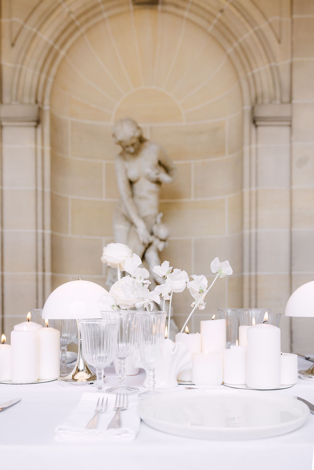 A white-clothed table with candles, glasses, plates and white flowers sits before a stone alcove holding a cherub statue.