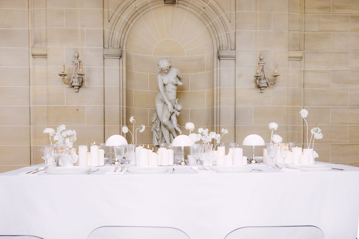 A long white-clothed table with candles and slender white flowers stands in front of a stone wall niche holding a statue.
