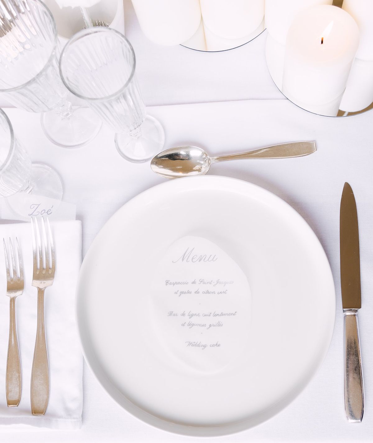 A white plate printed with a menu sits between silver cutlery, glasses and a lit candle, photographed from directly above on white linen.