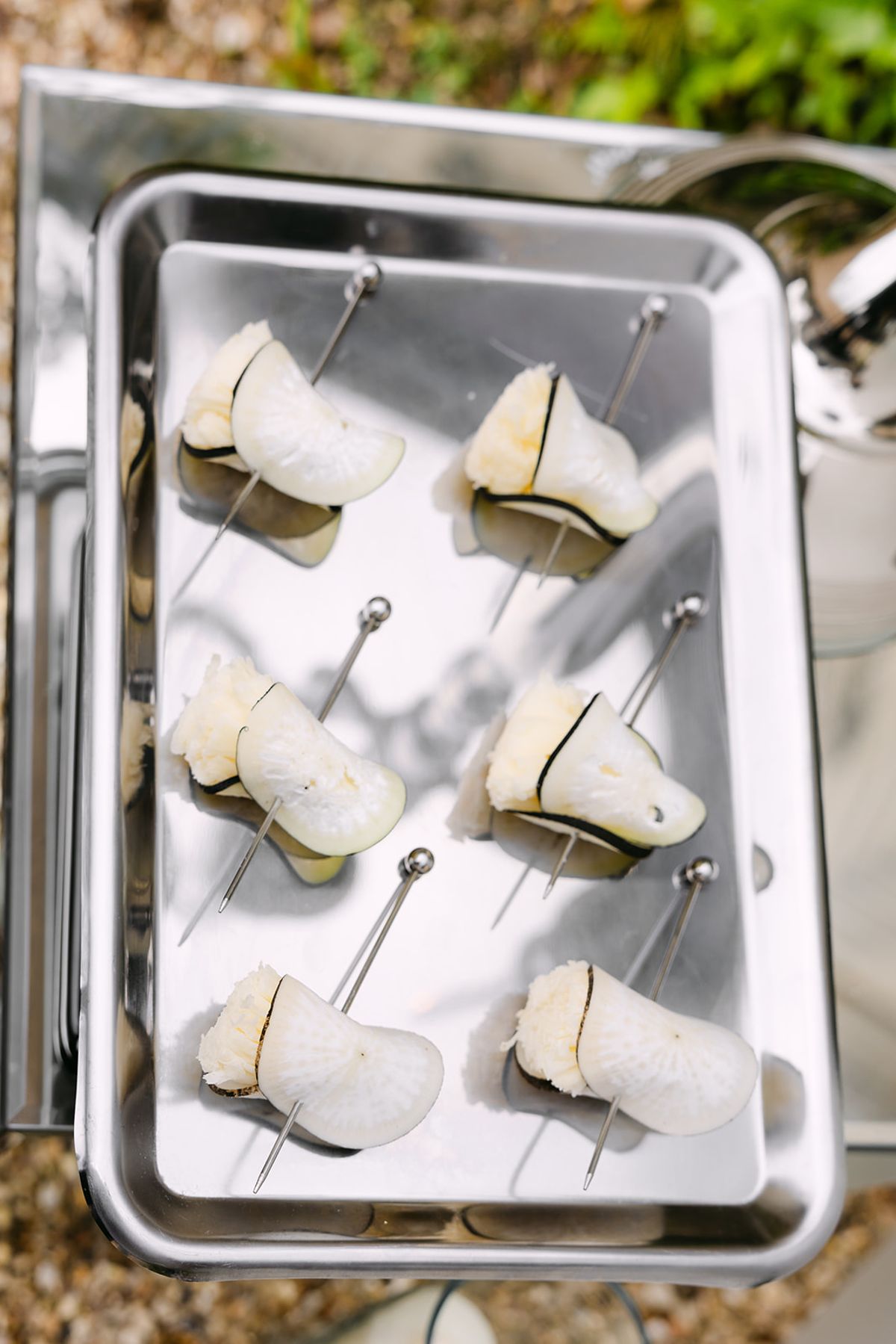 A stainless steel tray holds six small canapes on metal picks, set outdoors on gravel.