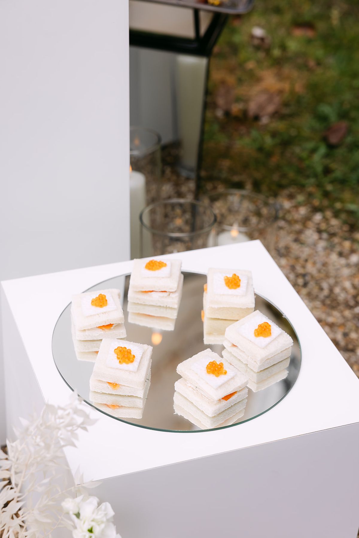 Small layered canapes topped with orange garnish sit on a mirrored round tray on a white plinth beside white flowers.