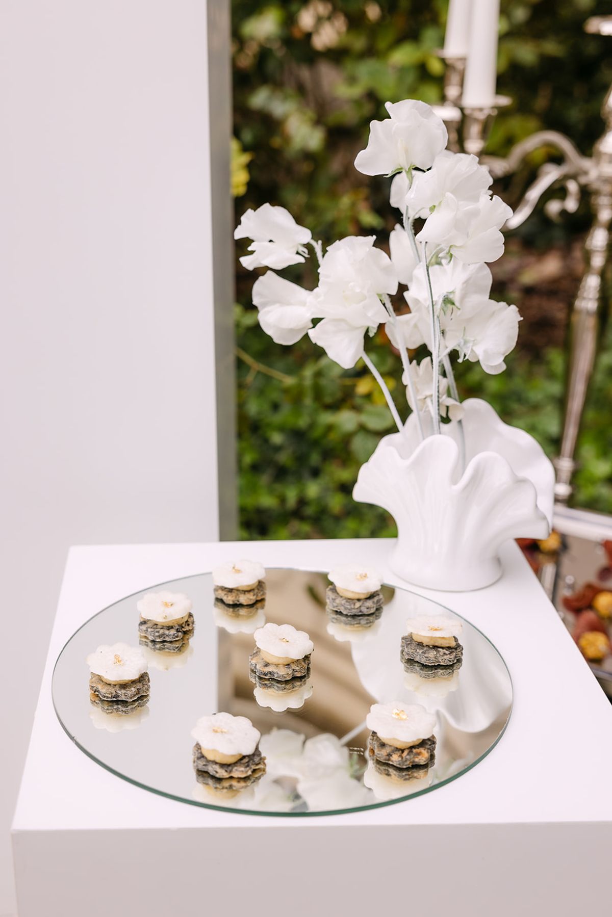 Small round desserts sit on a mirrored tray on a white plinth beside a white vase holding delicate white blossoms.