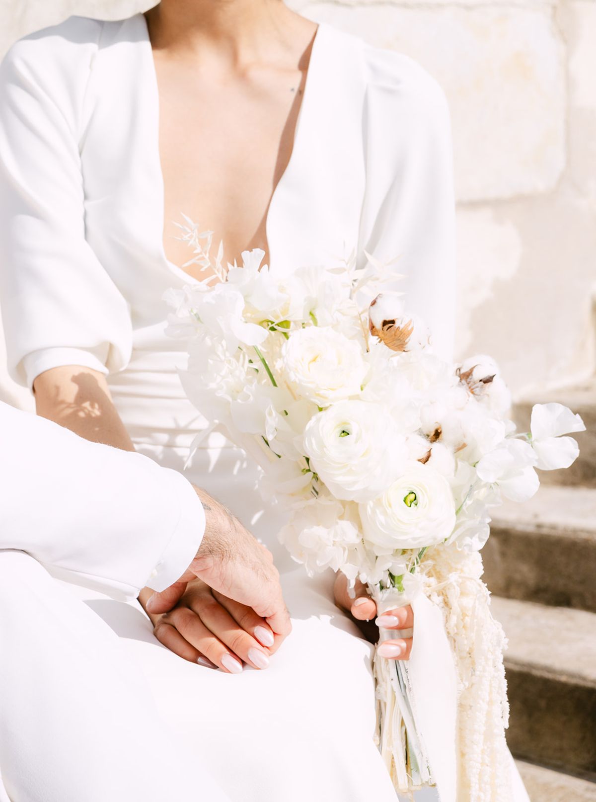 The groom's hand resting over the bride's as she holds a white bouquet of ranunculus, sweet peas and cotton