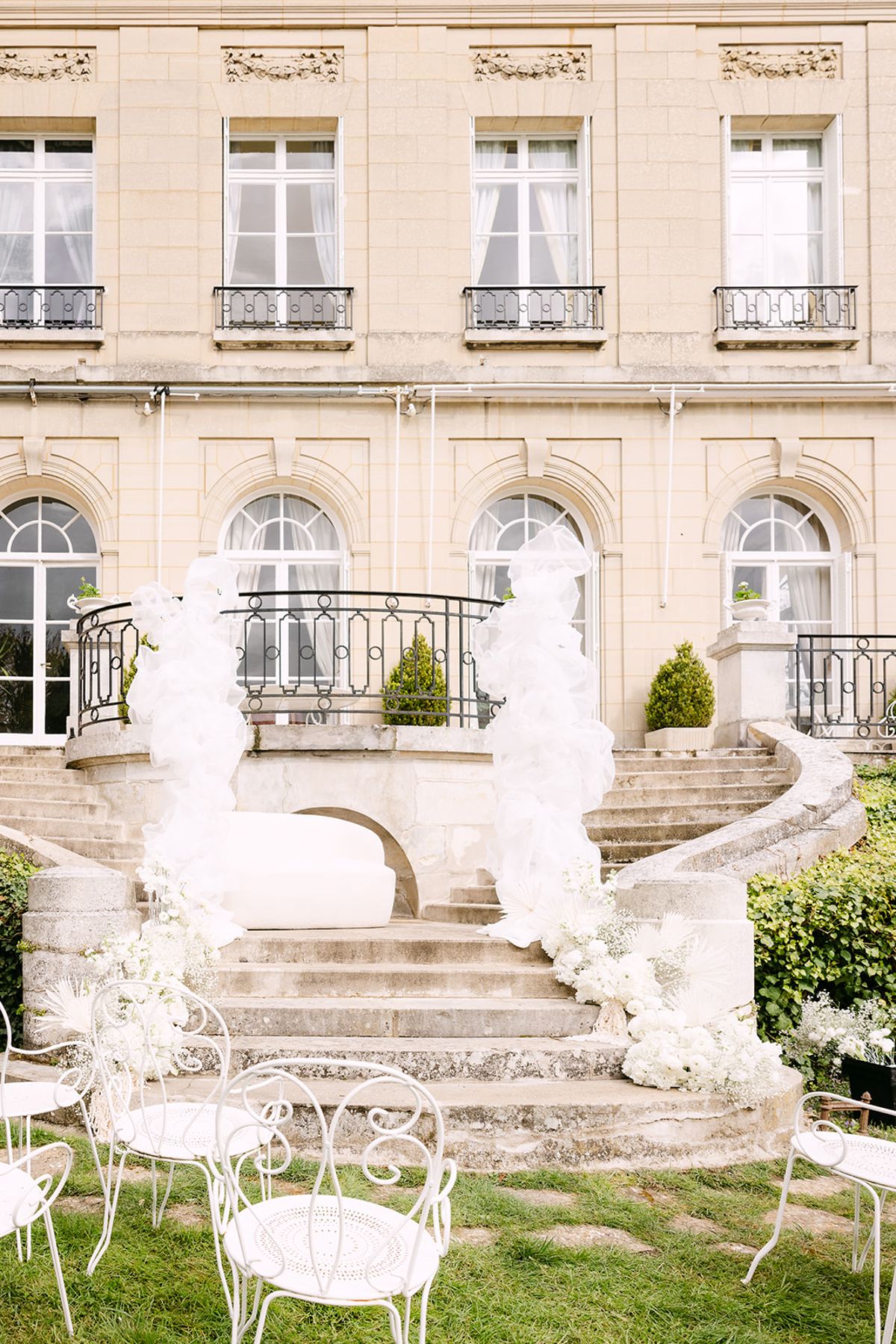 A cream stone chateau with tall windows, a curved staircase, white tulle sculptures and white chairs on the lawn below.