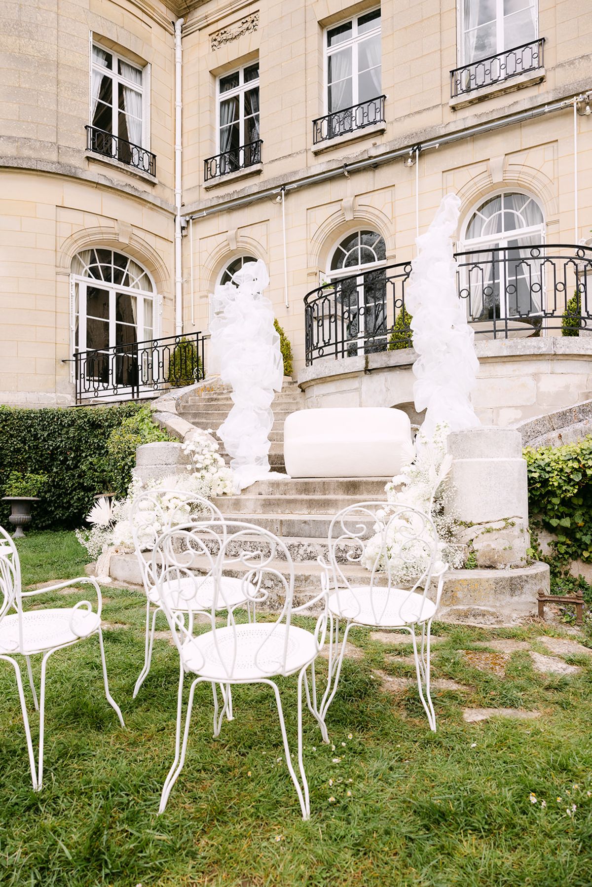 A stone chateau facade overlooks a lawn with white metal chairs and tall white floral and tulle arrangements beside a curved staircase.