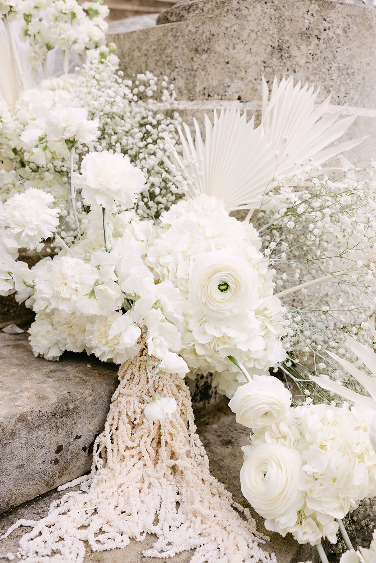 A cluster of white roses, ranunculus, dried palm and pearl strands spills over weathered stone steps.