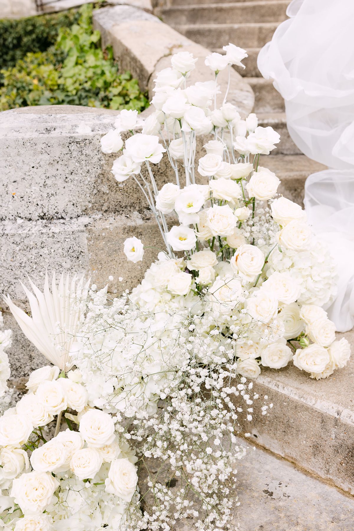 White roses, baby's breath and dried palm leaves are arranged on weathered stone steps beside soft white tulle.