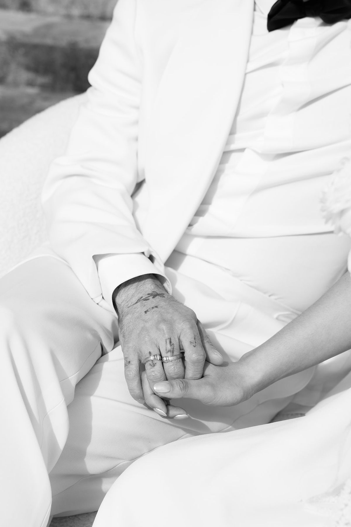 A black and white close-up of two hands clasped together on the lap of a groom in a white suit, a ring visible.
