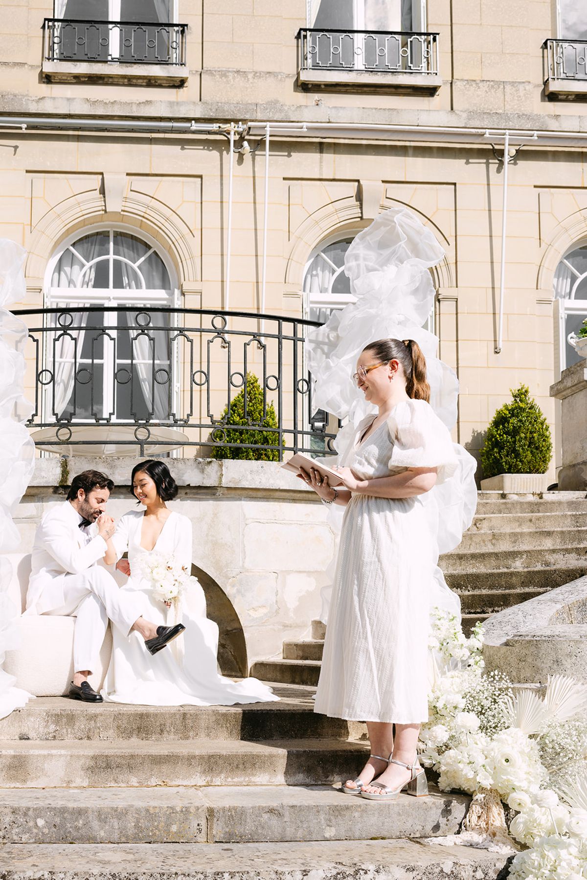 Groom kissing the bride's hand as they sit on the terrace steps while a woman in a white dress reads from a book