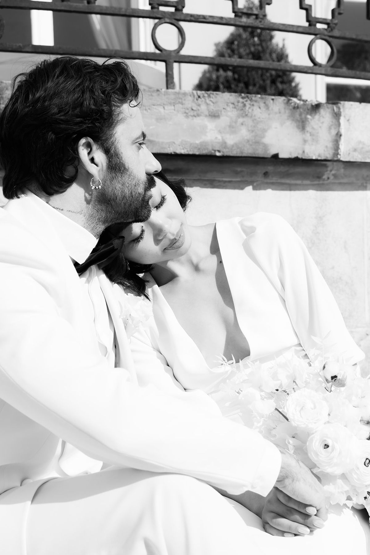 In black and white, the groom sits with the bride resting her head on his shoulder, holding a bouquet near a balustrade.