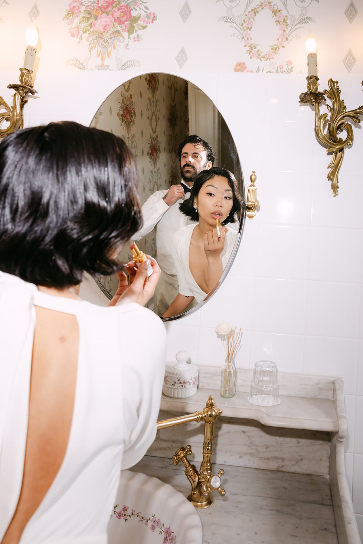 The bride with a short dark bob applies lipstick in an oval mirror, while the groom is reflected behind her in a bathroom.
