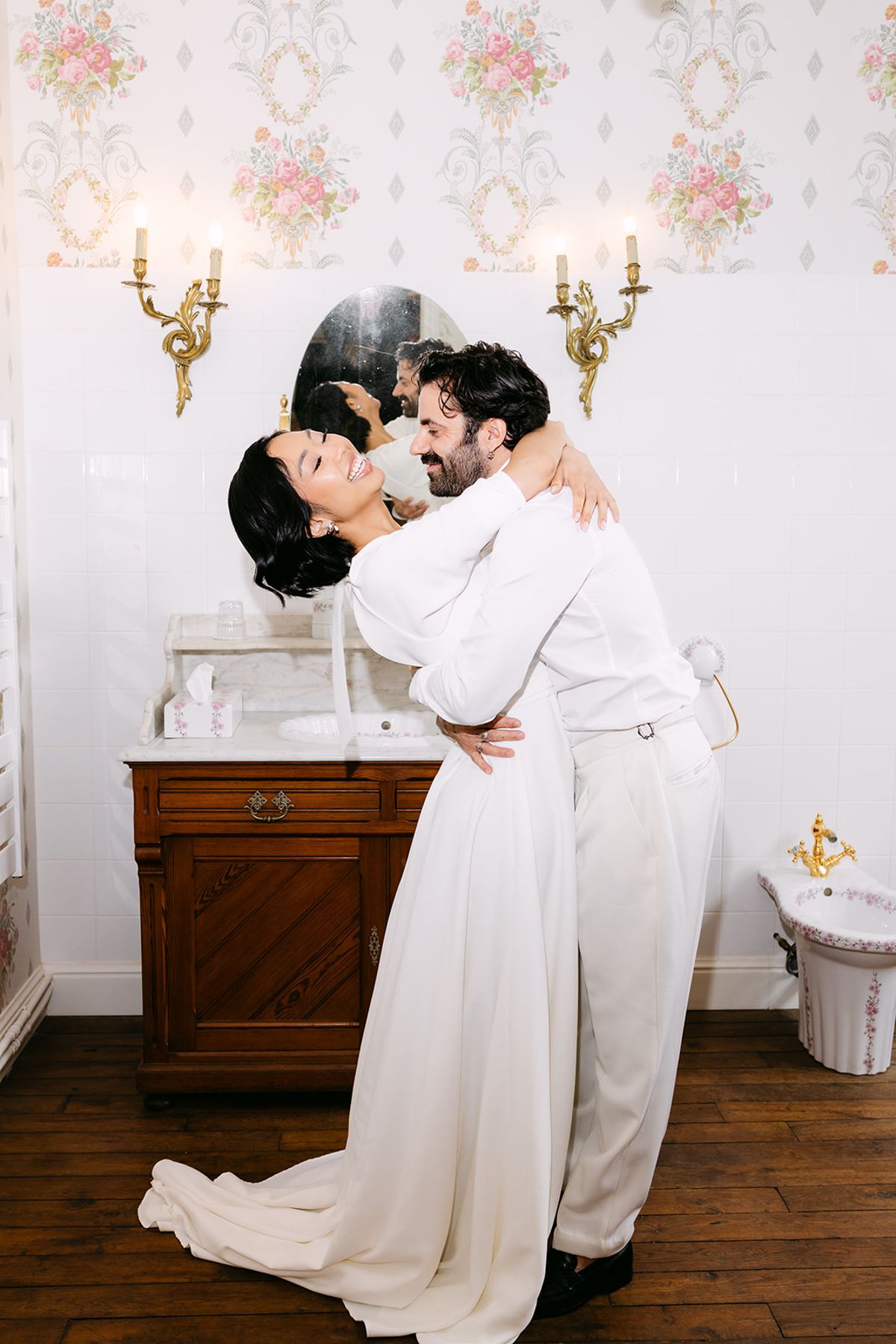 Bride and groom embracing beside a wooden washstand in a rose-papered bathroom at Château du Prieuré d'Évecquemont