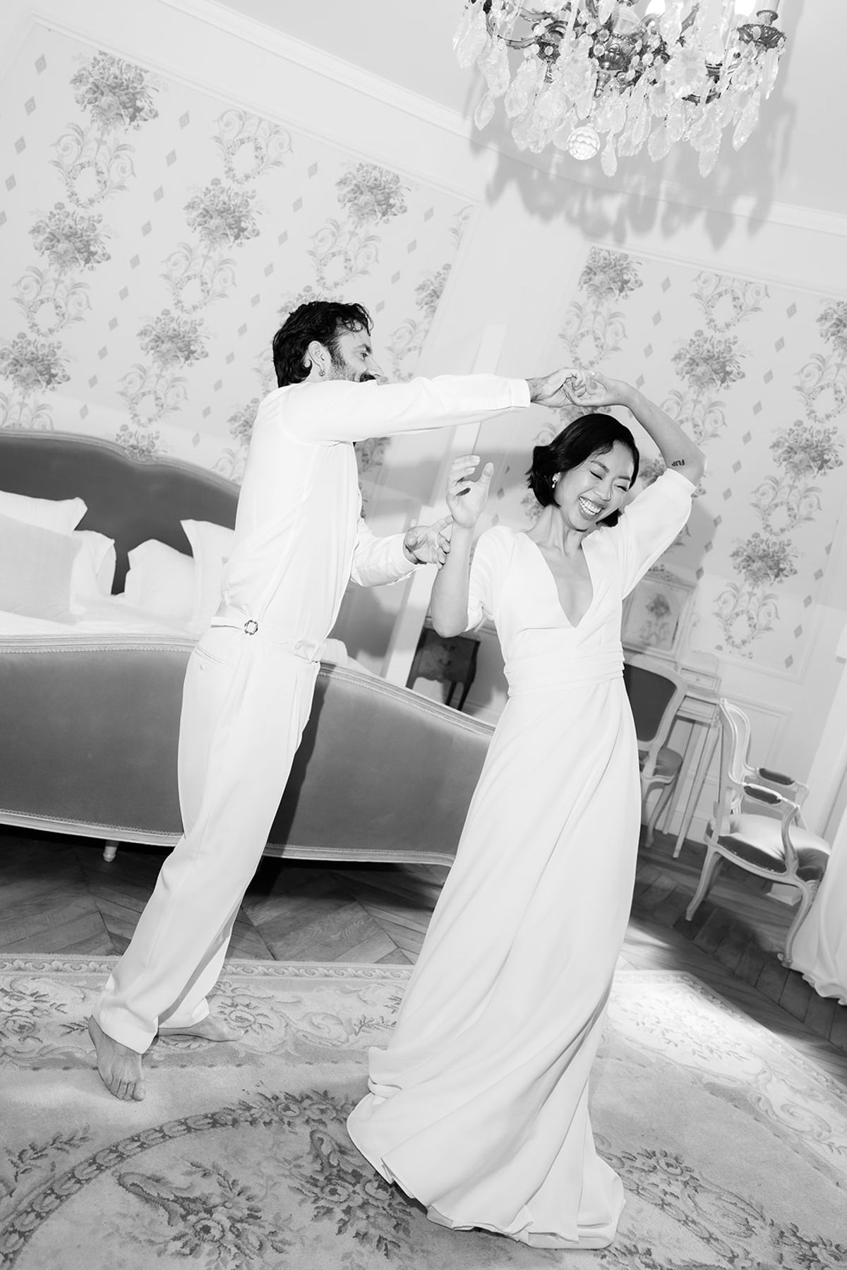 In black and white, the groom and bride dance playfully in a room with floral wallpaper, a sofa and a chandelier.