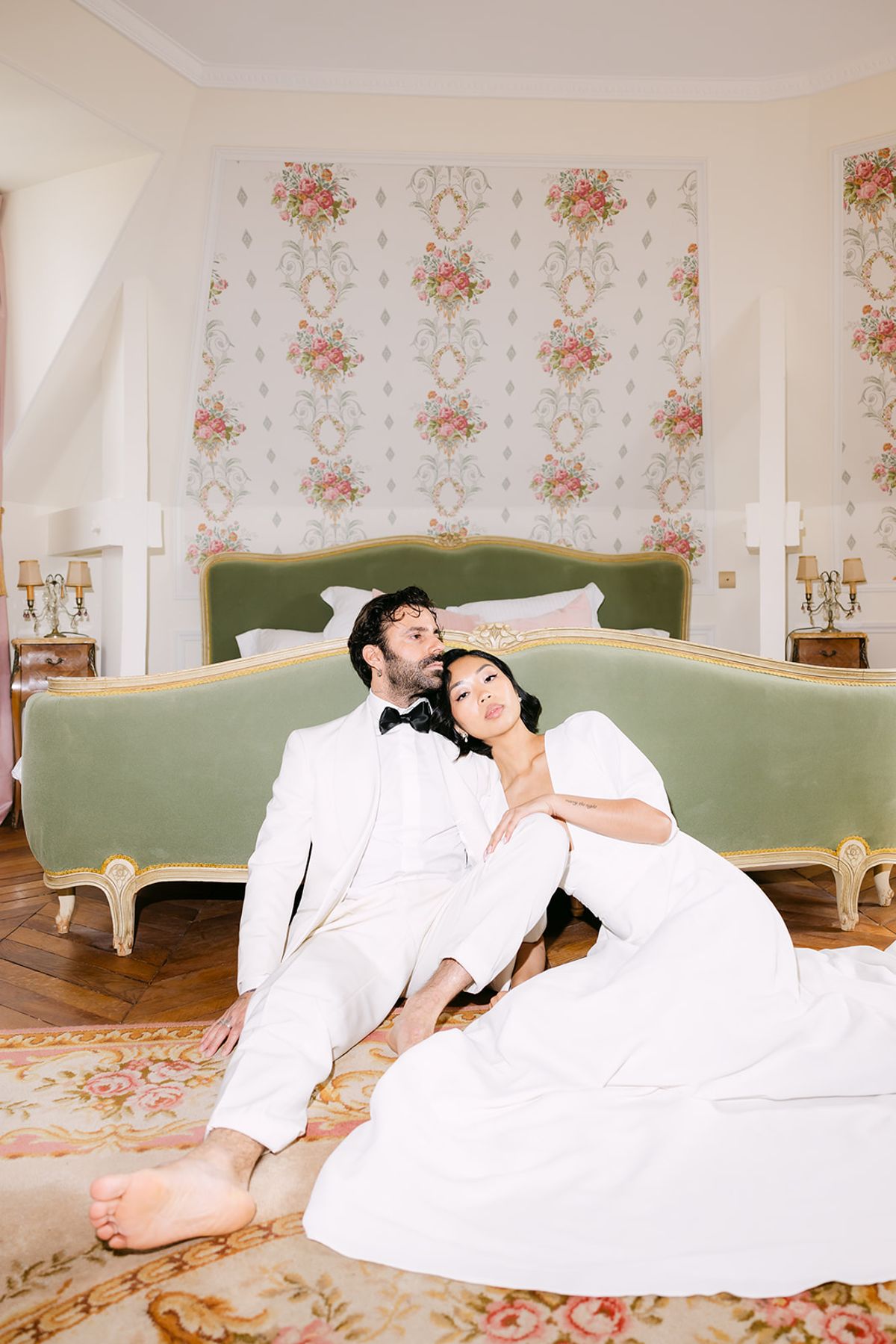The couple in white recline together on a patterned rug in front of a green upholstered bed with floral wallpaper behind.