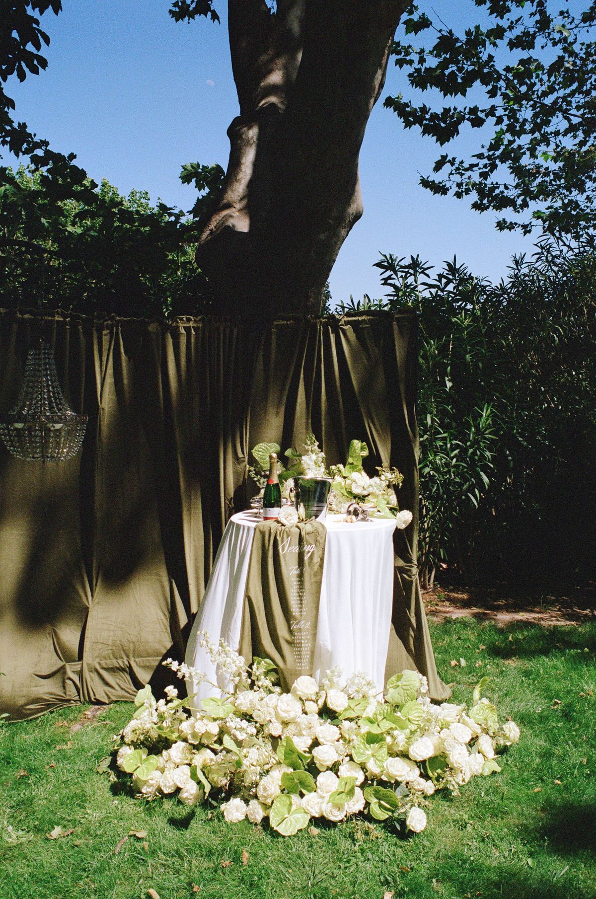 Cocktail table dressed in olive linen with a single floral stem beneath the trees at dusk