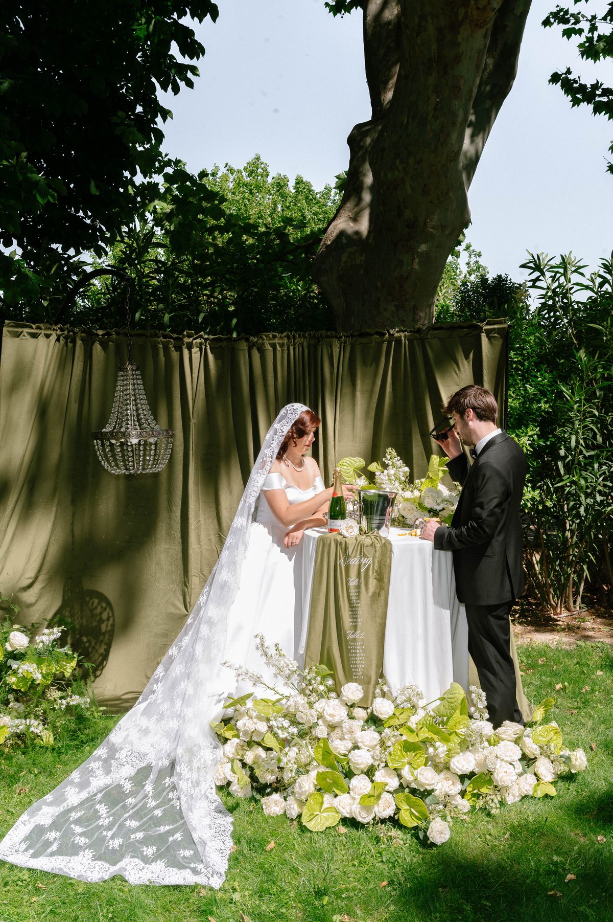 Couple seated together at their reception table beneath string-lit trees at dusk