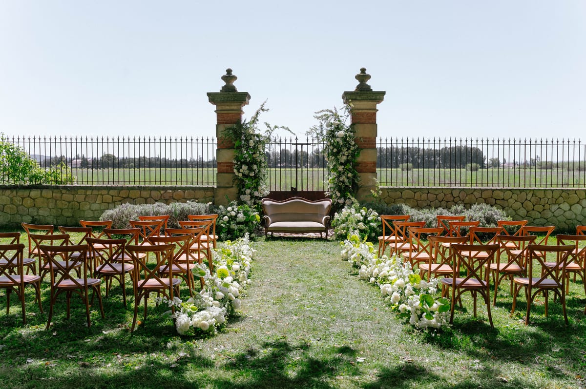 The ceremony aisle set with wooden chairs leading to the floral stone archway under open sky