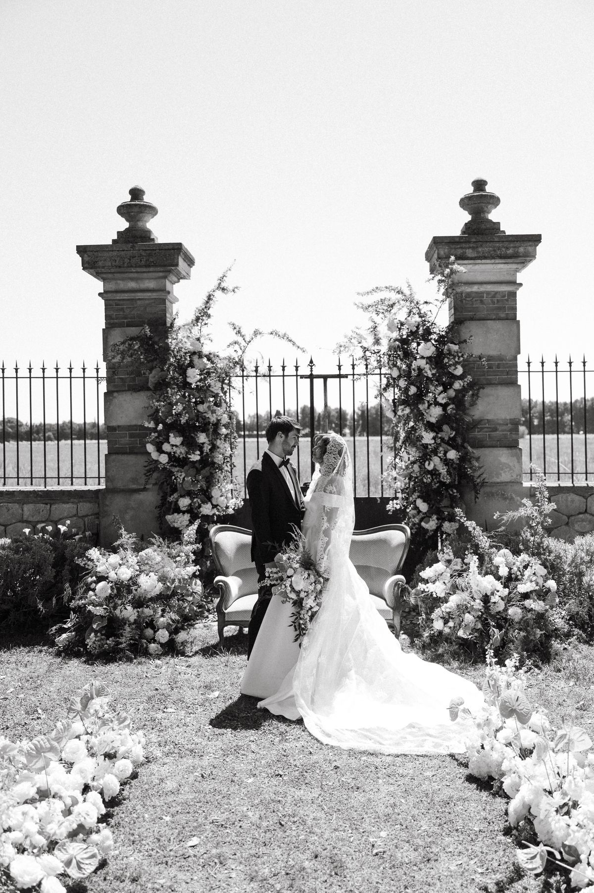 Black-and-white image of the couple walking together through the stone pillar entrance, floral garlands overhead