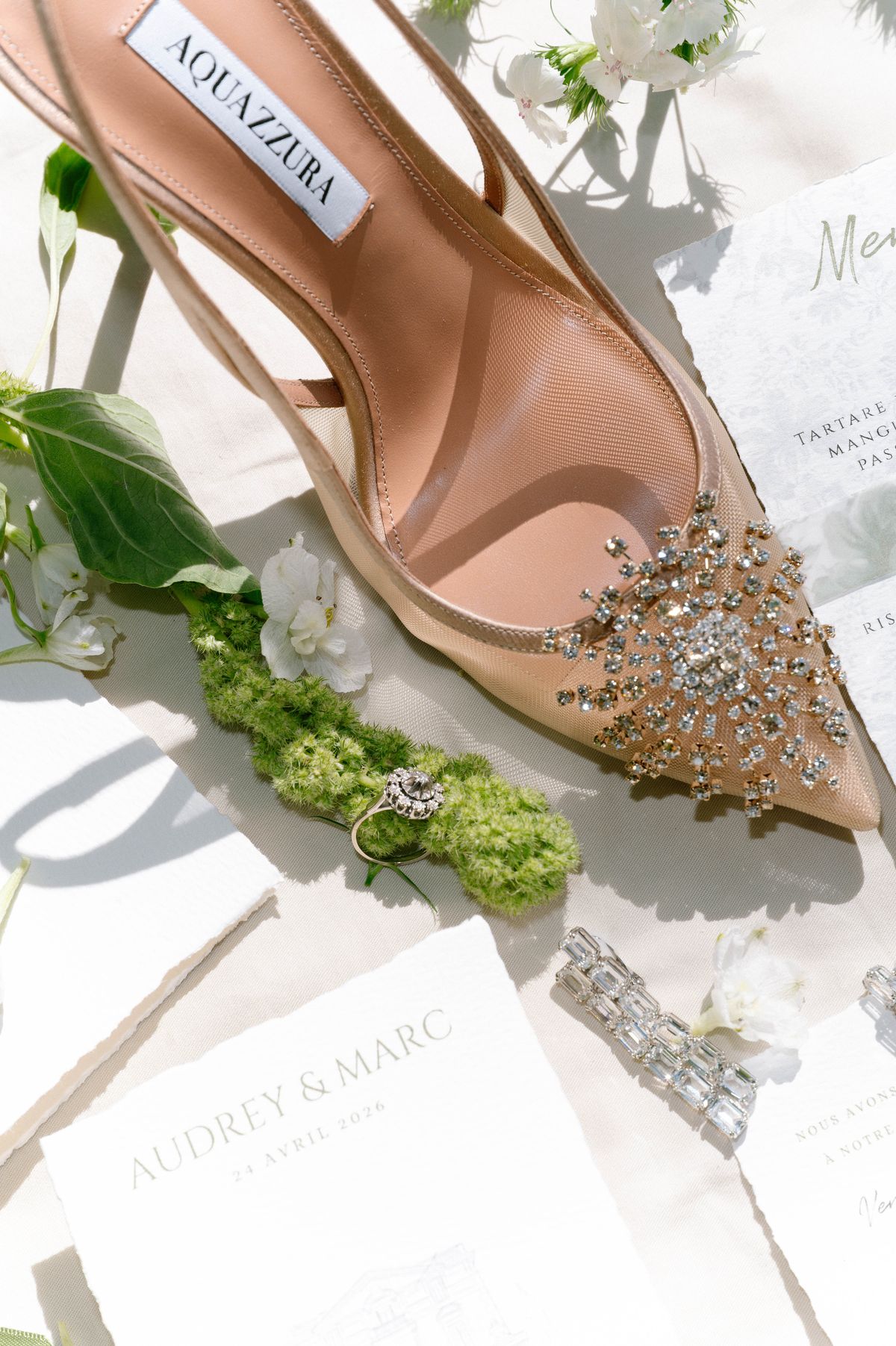 Crystal-embellished heel resting on a bed of white flowers and greenery beside a printed invitation