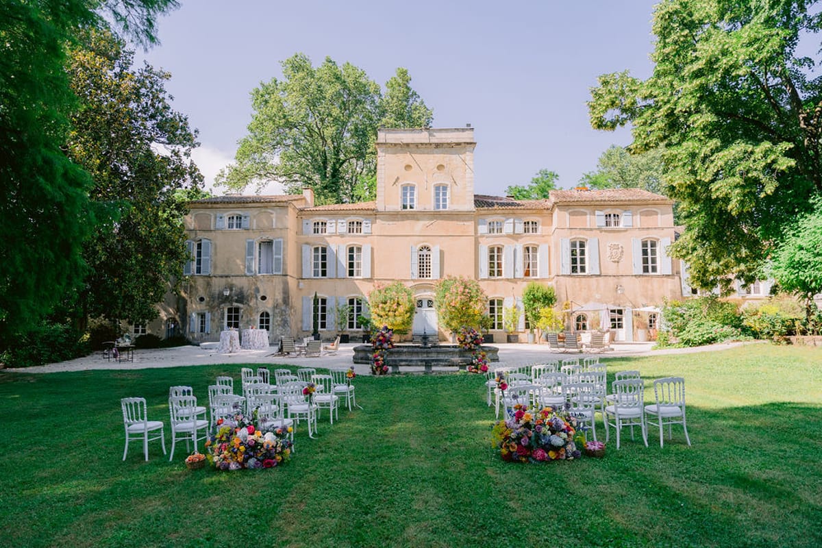 A large stone chateau stands behind a lawn where white chairs line an aisle that ends in a colourful flower arrangement.