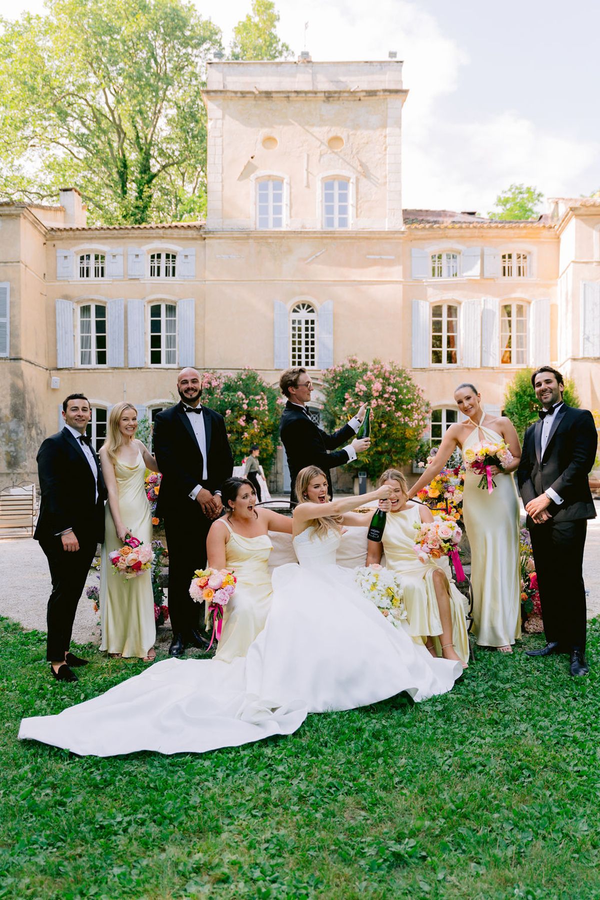 The bride sits on the grass in her gown, surrounded by smiling bridesmaids and groomsmen, with a pale chateau behind them.