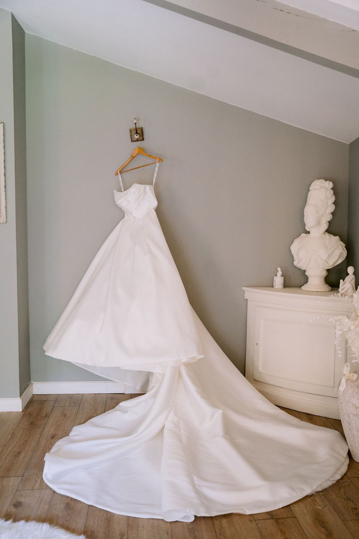 A white wedding gown with a long train hangs from a hook in a pale room with a classical bust and a white cabinet.