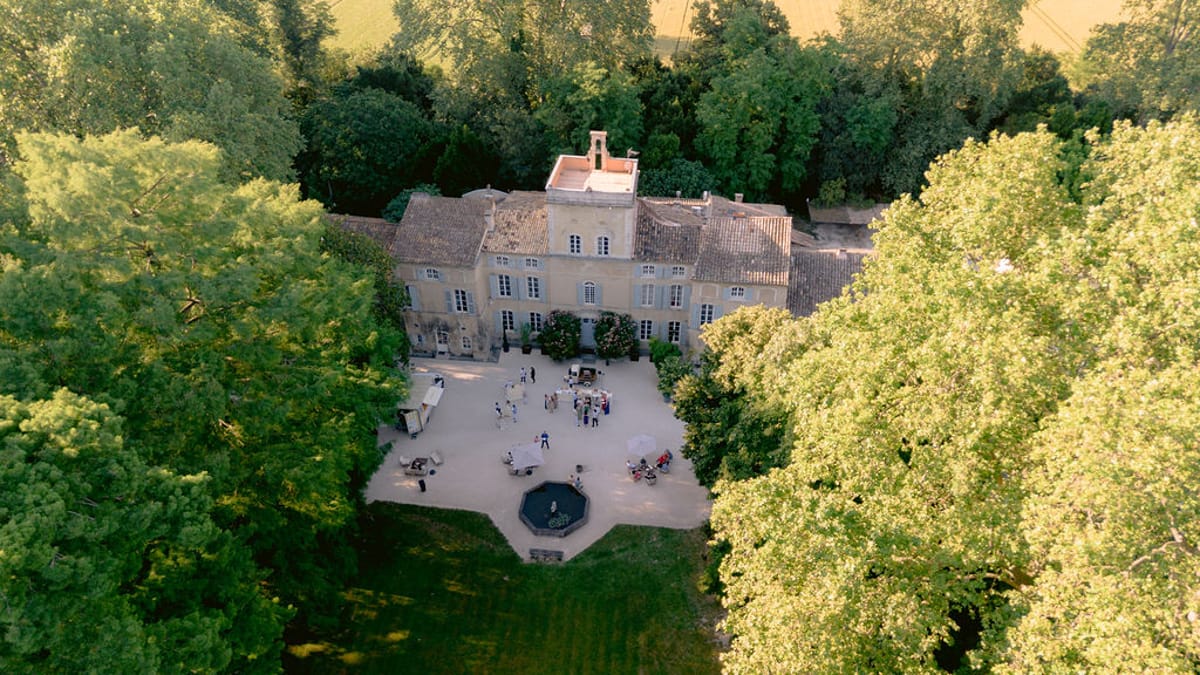 An aerial view shows a stone chateau and its gravel courtyard surrounded by dense green trees lit by warm light.