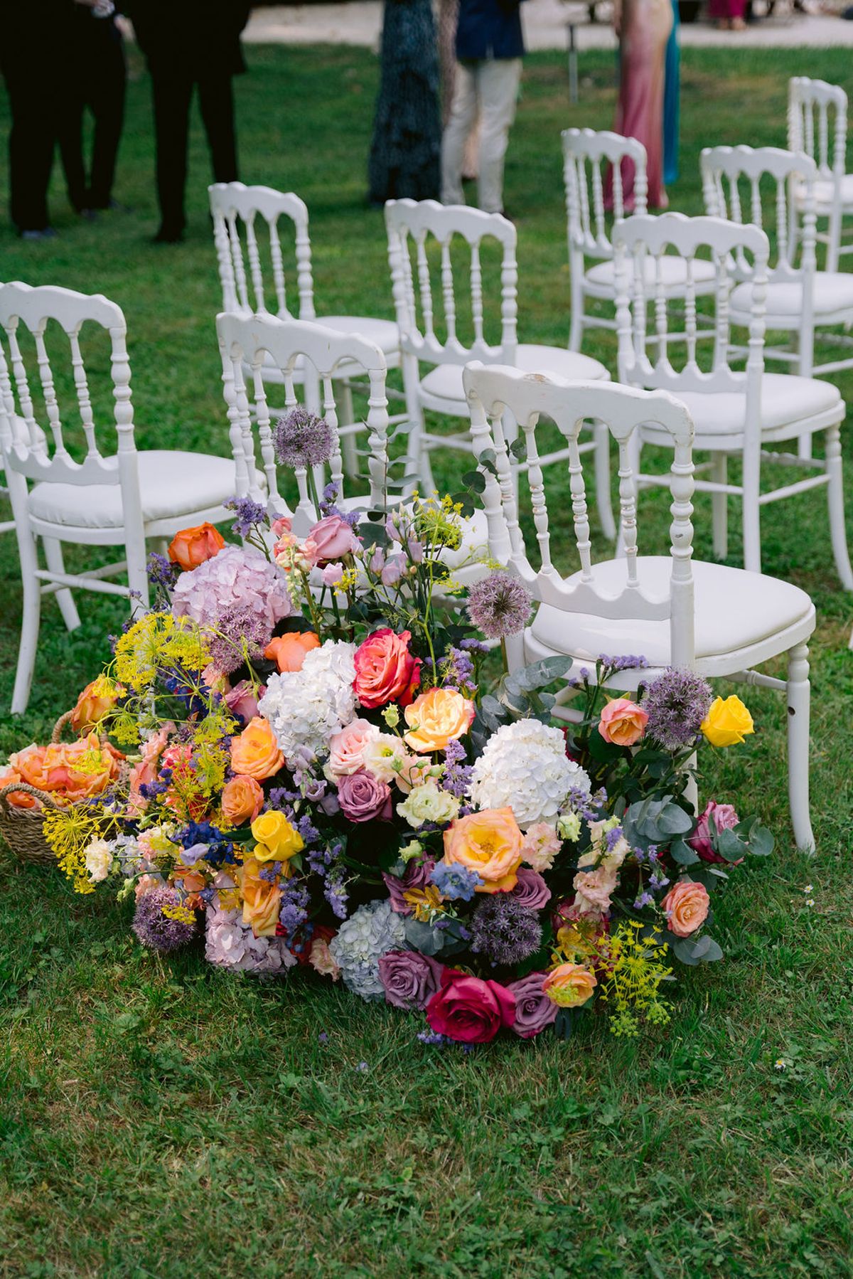 White chairs stand in rows on a lawn beside a large ground arrangement of orange, pink, purple and white flowers.