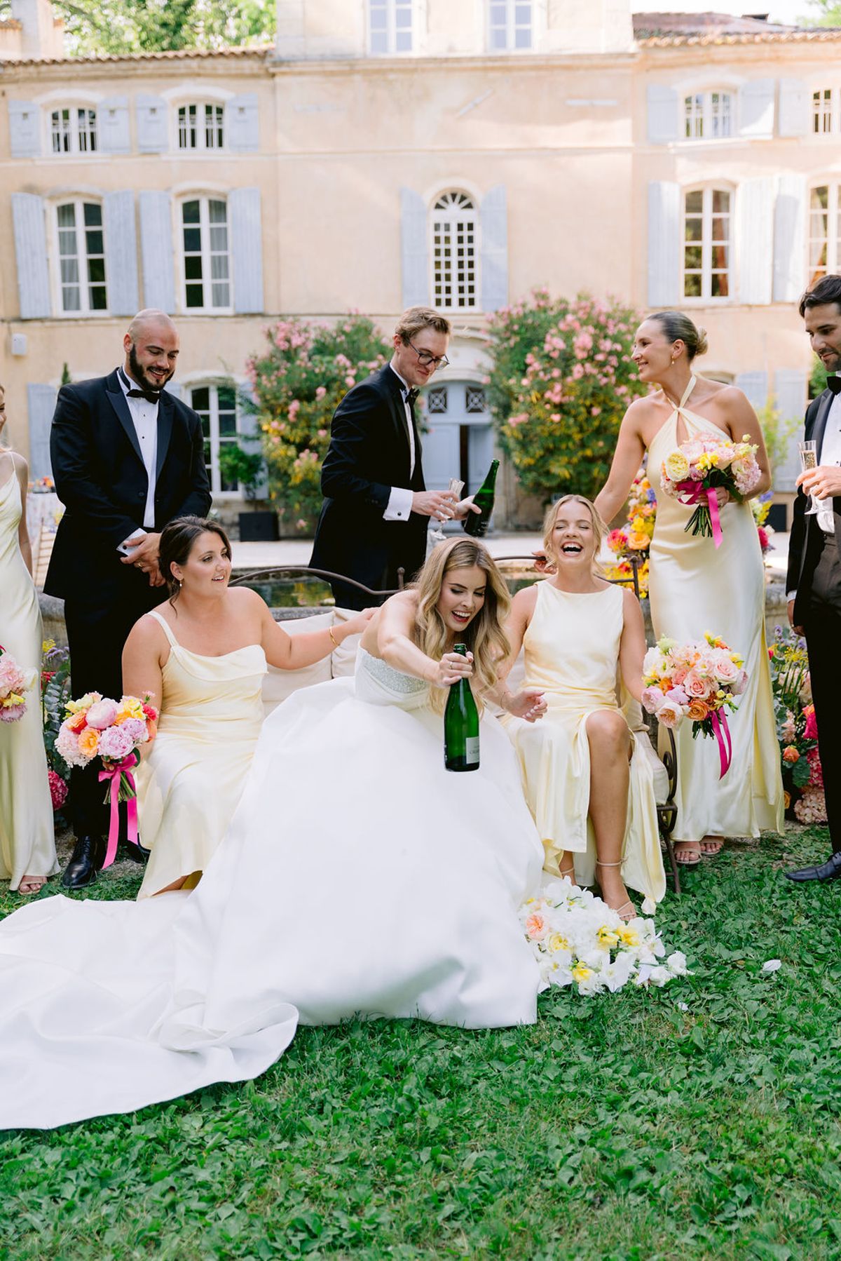 The bride sits on the grass in her gown laughing with bridesmaids and men in tuxedos, one holding a champagne bottle, before a chateau.