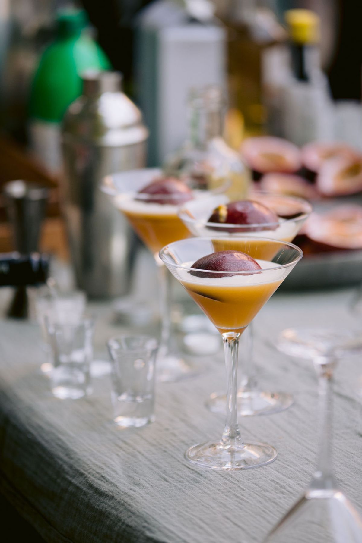 Several orange cocktails in stemmed glasses stand on a table beside a metal cocktail shaker and small shot glasses.