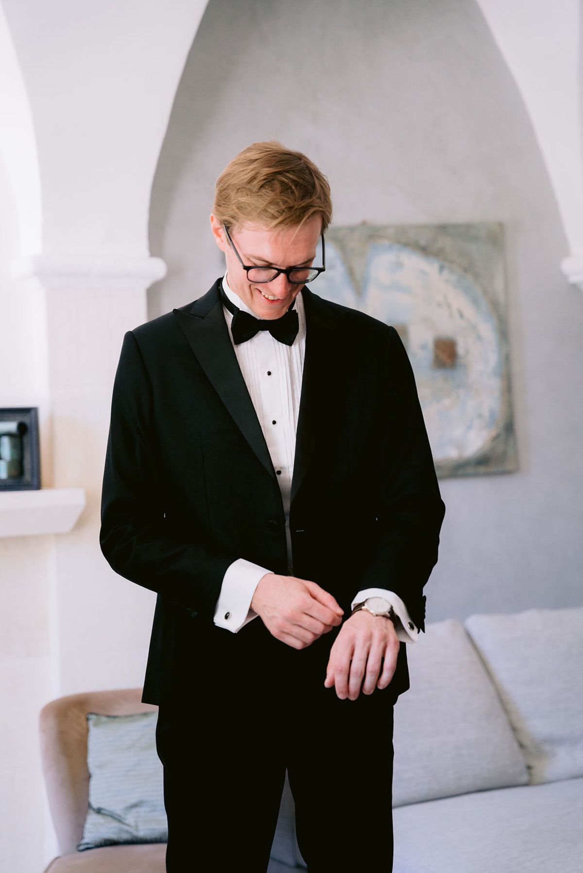 A man with glasses, dressed in a black tuxedo and bow tie, looks down while adjusting his shirt cuff in a bright room.