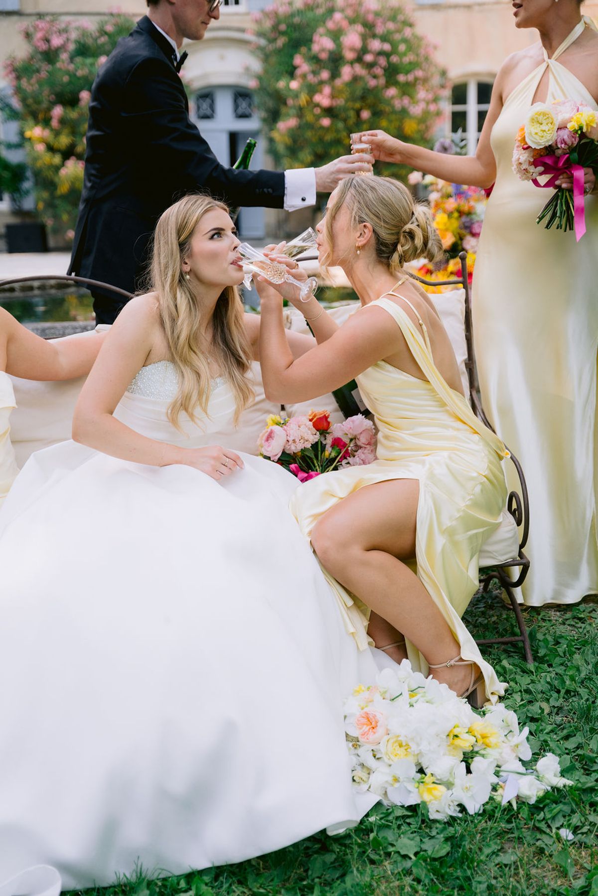 The bride in a white gown sits on the grass drinking champagne with a bridesmaid in yellow, while others stand around them.