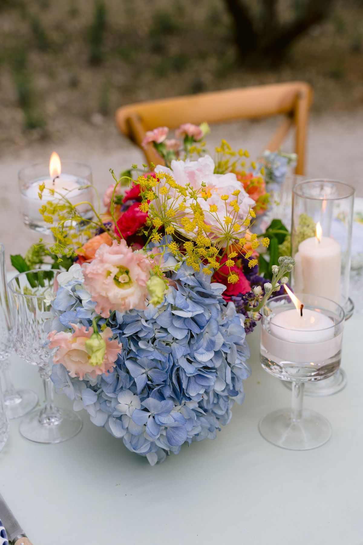 A centrepiece of blue hydrangeas and bright red, orange and yellow flowers sits beside lit candles and crystal glasses on a pale table.
