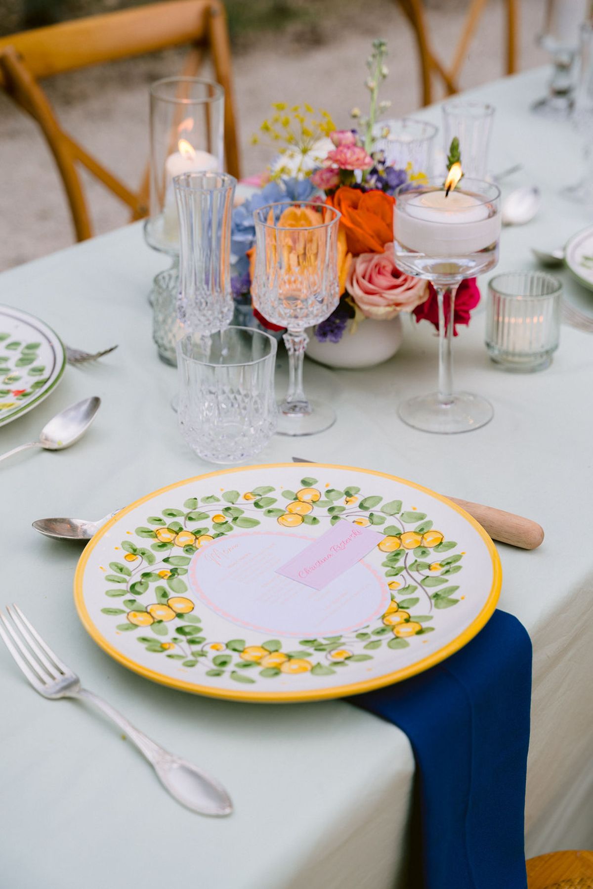 A table setting with a hand-painted plate, navy napkin, crystal glasses, candles and a colourful floral centrepiece on a pale green tablecloth.