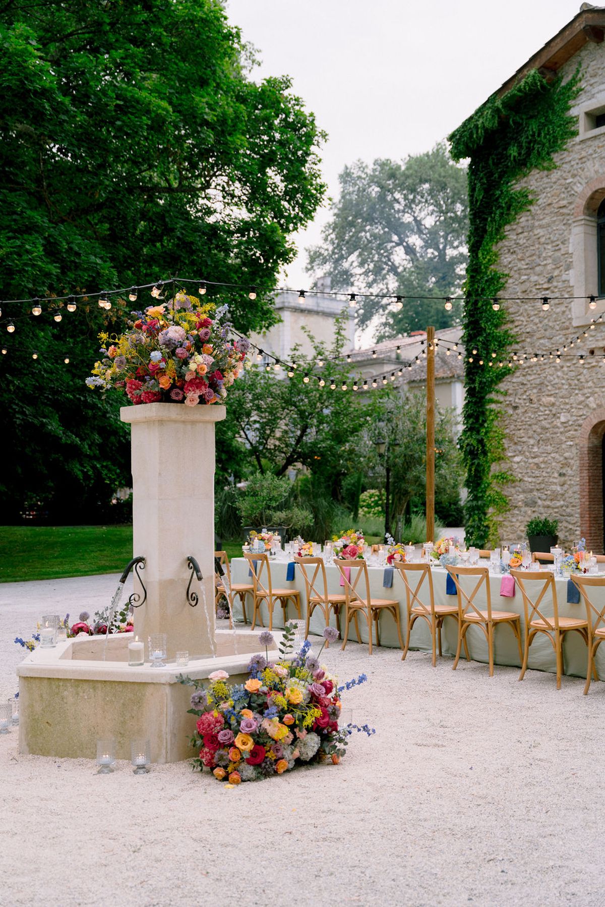 A stone fountain topped with bright flowers stands in a gravel courtyard beside long tables under festoon lights.